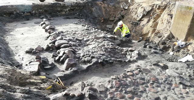 En arkeolog i skyddskläder står i en stor grop där stenläggning grävts fram.