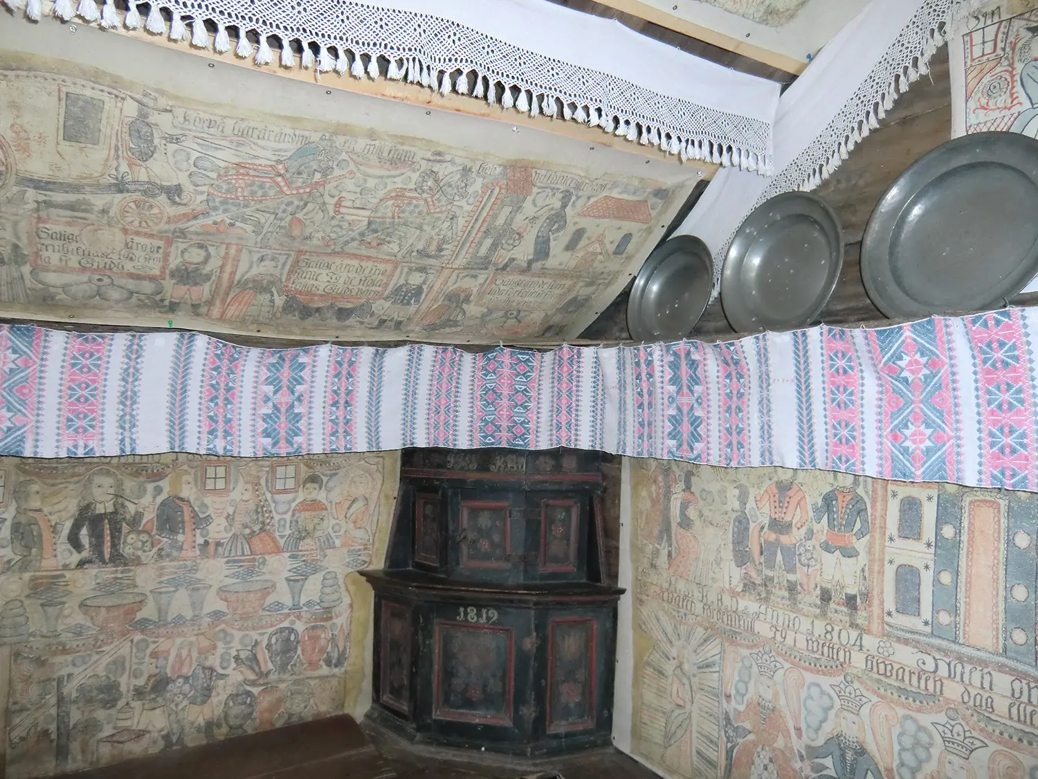 Interiör från den historiska byggnaden Bollaltebygget med opphämtaväv och bonadsmålningar som utgår från ett hörn och fortsätter på båda sidor längs väggen.