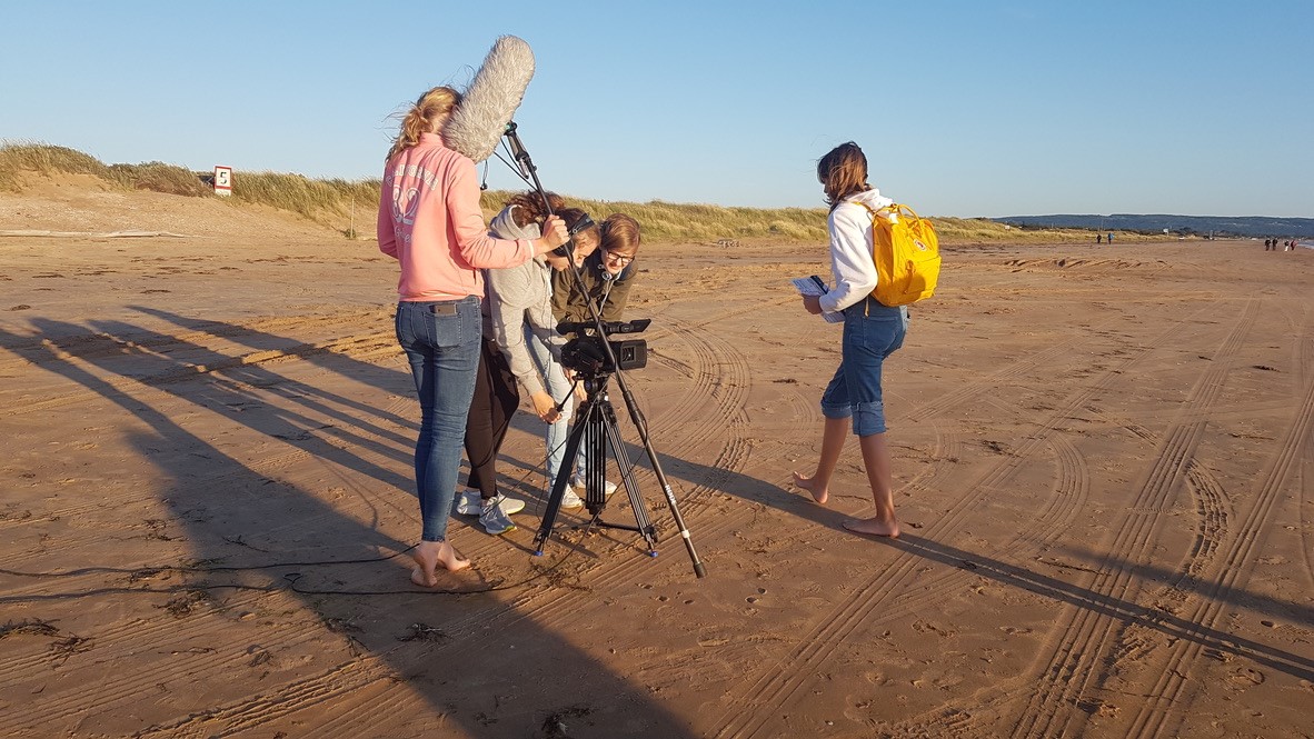 Fyra tjejer spelar in film på en sandstrand.