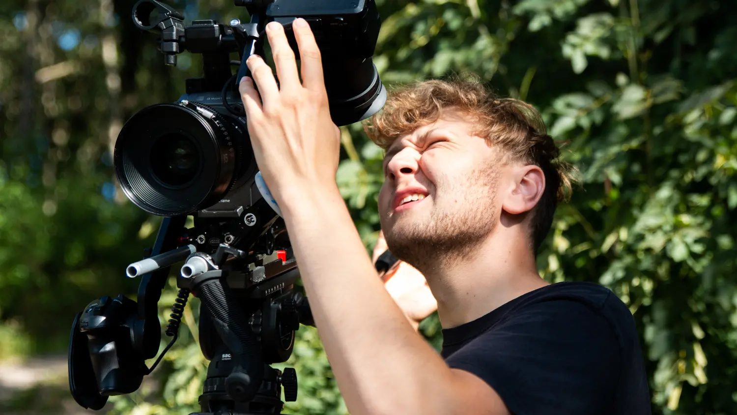 En person står ute i grönskan och kisar mot ljuset, samtidigt som han håller i och tittar på en filmkamera på stativ.