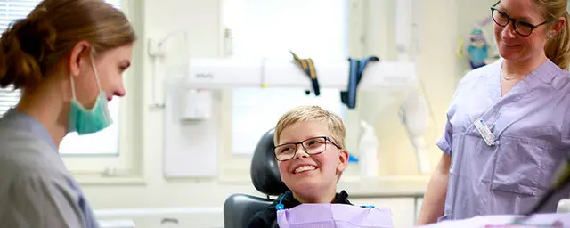 Pojke med glasögon sitter i en tandläkarstol, kvinnlig tandläkare och kvinnlig tandsköterska står på var sin sida om honom.