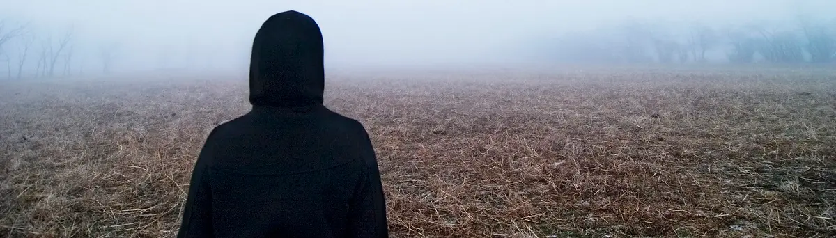 Person i svart luvtröja tittar ut över ett dimmigt fält.