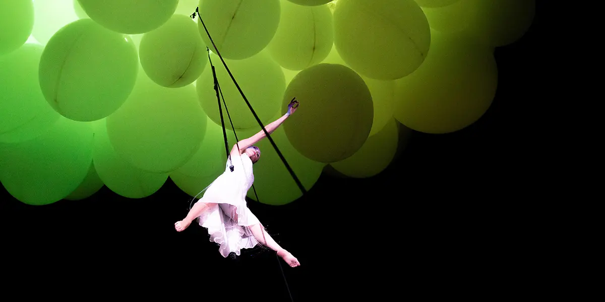 En akrobat hänger i linor under en massa ljusgröna ballonger.