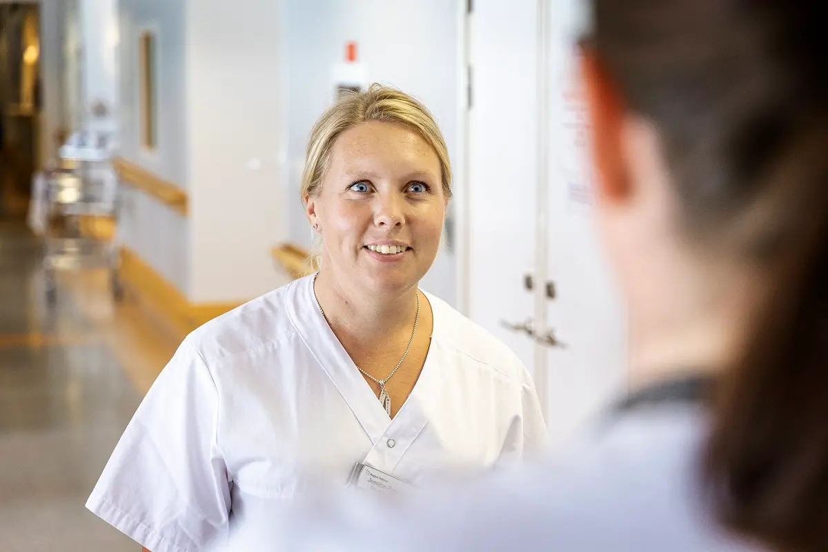 Blond kvinna iförd vita vårdkläder, i en sjukhuskorridor.
