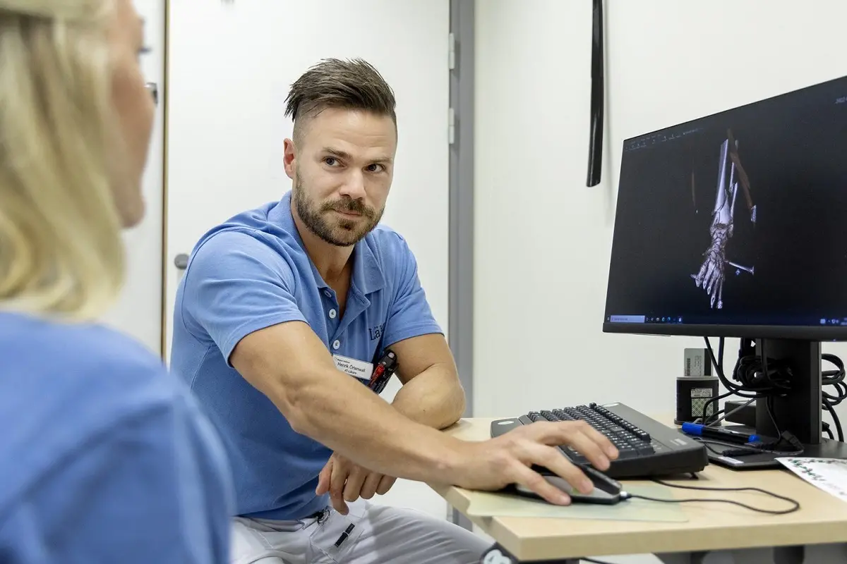 Man med mörkt hår och skägg sitter vid ett skrivbord med en datorskärm som visar en röntgenbild, han har blå vårdkläder på sig.