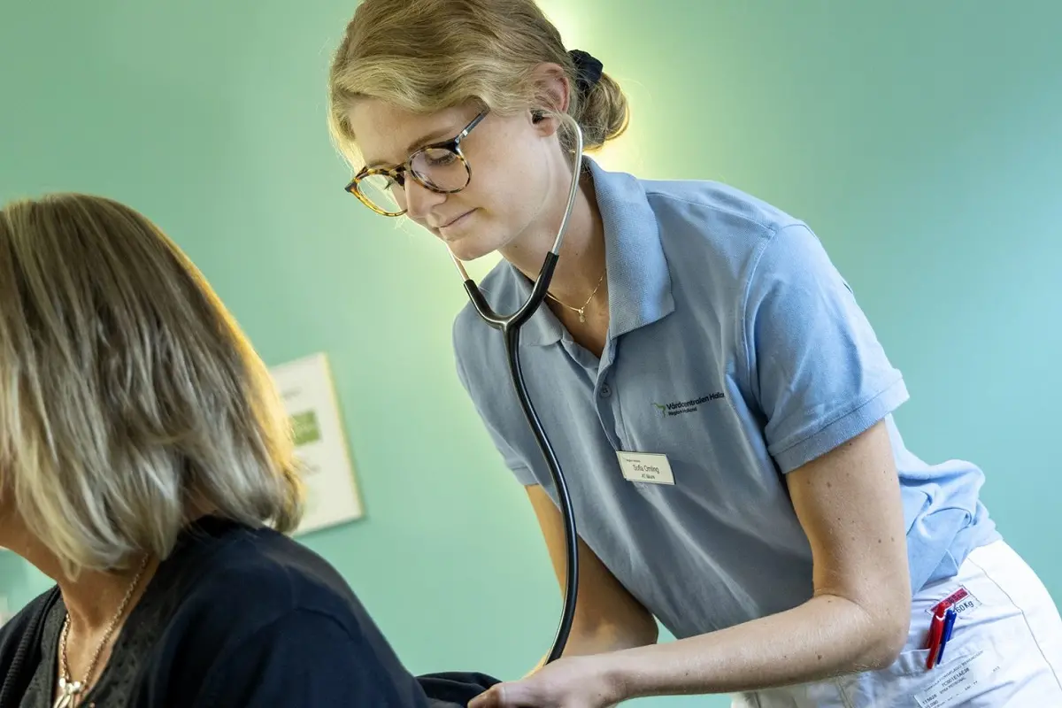Blond kvinna med glasögon iförd vårdkläder håller ett stetoskop mot en persons rygg.