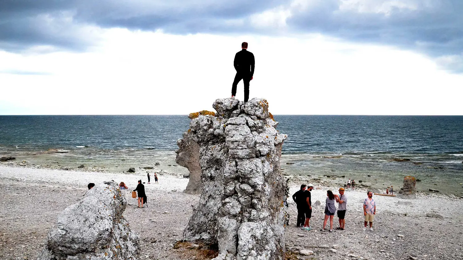 En person står högst upp på en stor rauk på en kalkvit strand vid havet.
