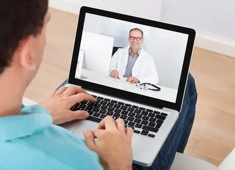 Man med laptop i knät, en läkare syns på datorns skärm.