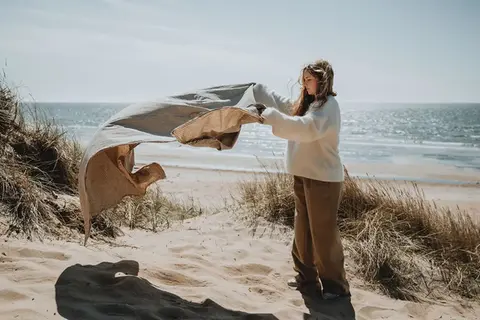 Tjej med långt hår breder ut en filt på stranden. I bakgrunden hav och vass.