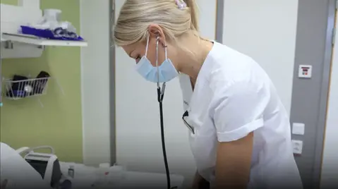 Blond kvinna i vårdkläder och munskydd använder ett stetoskop.