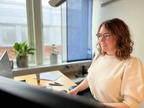 En brunhårig kvinna med glasögon sitter vid ett skrivbord framför en datorskärm.