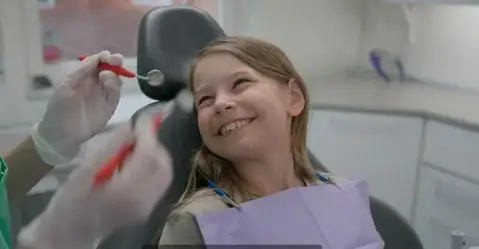 En leende flicka sitter i en tandläkarstol, en tandläkare håller upp verktyg för tandvård.